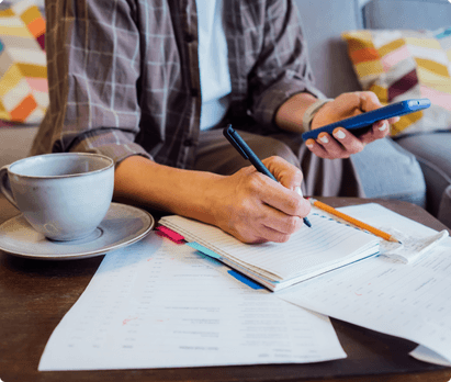 A person writing in a notebook while holder a phone