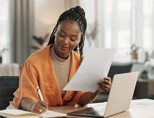 A woman sitting at a desk holding a piece of paper and writing in a notebook