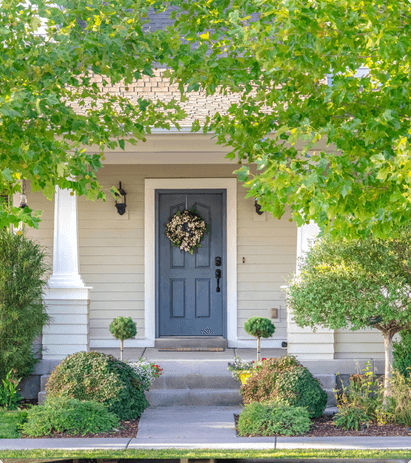 A beige house with trees and shrubs in front of it